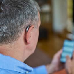 Close Up Of a Man Wearing Wireless Hearing Aid Using Mobile Phone At Home